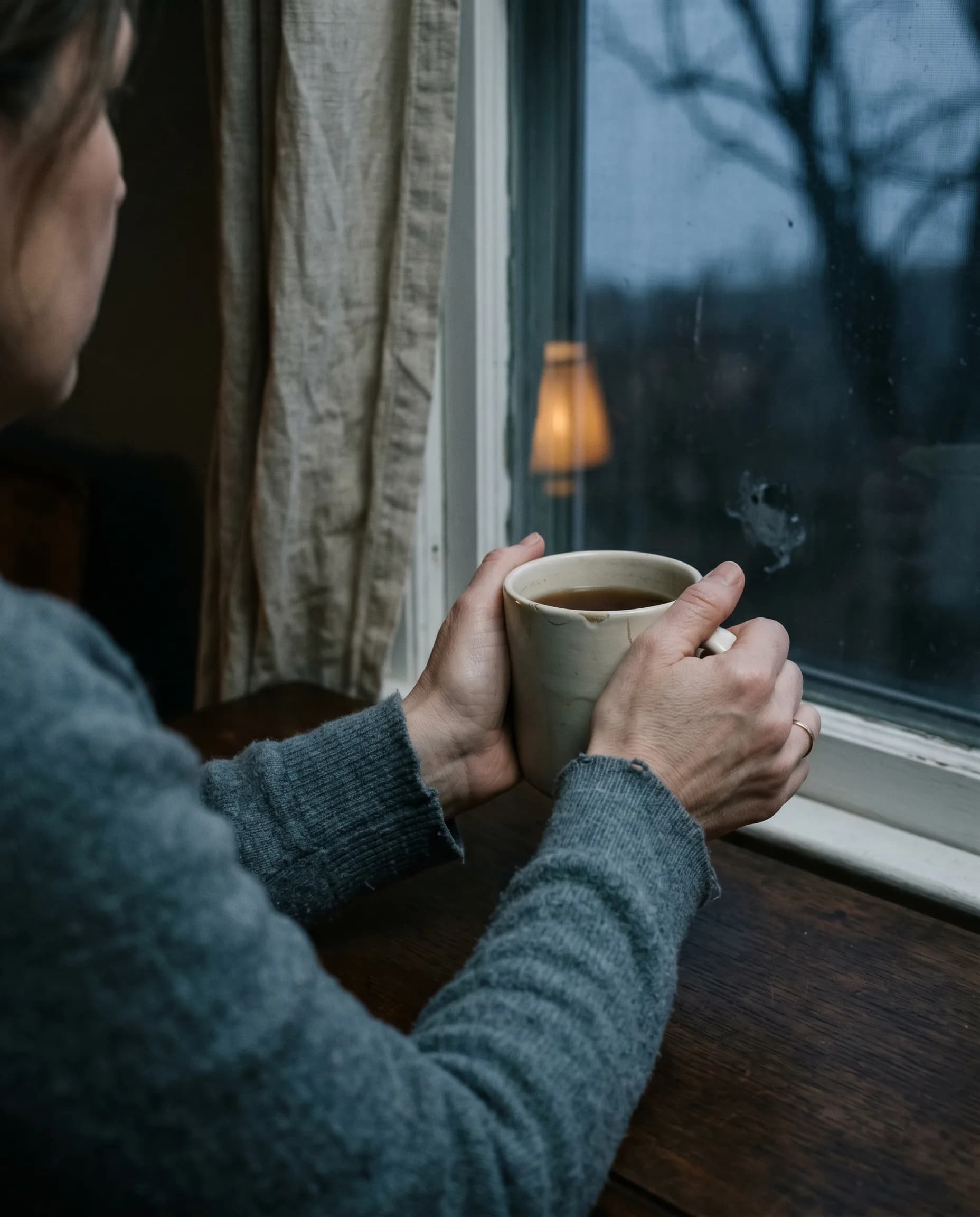 A woman's hands wrap around a ceramic mug of tea, held by an overcast morning window.